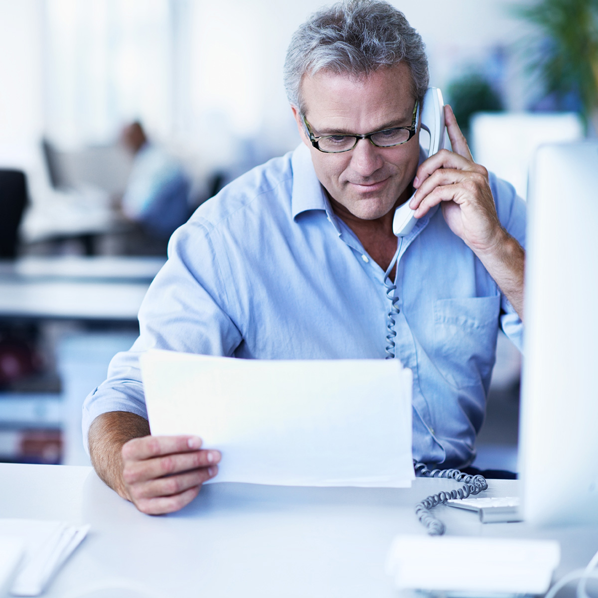 Man in office at his desk holding a piece of paper with desktop computer in front of him